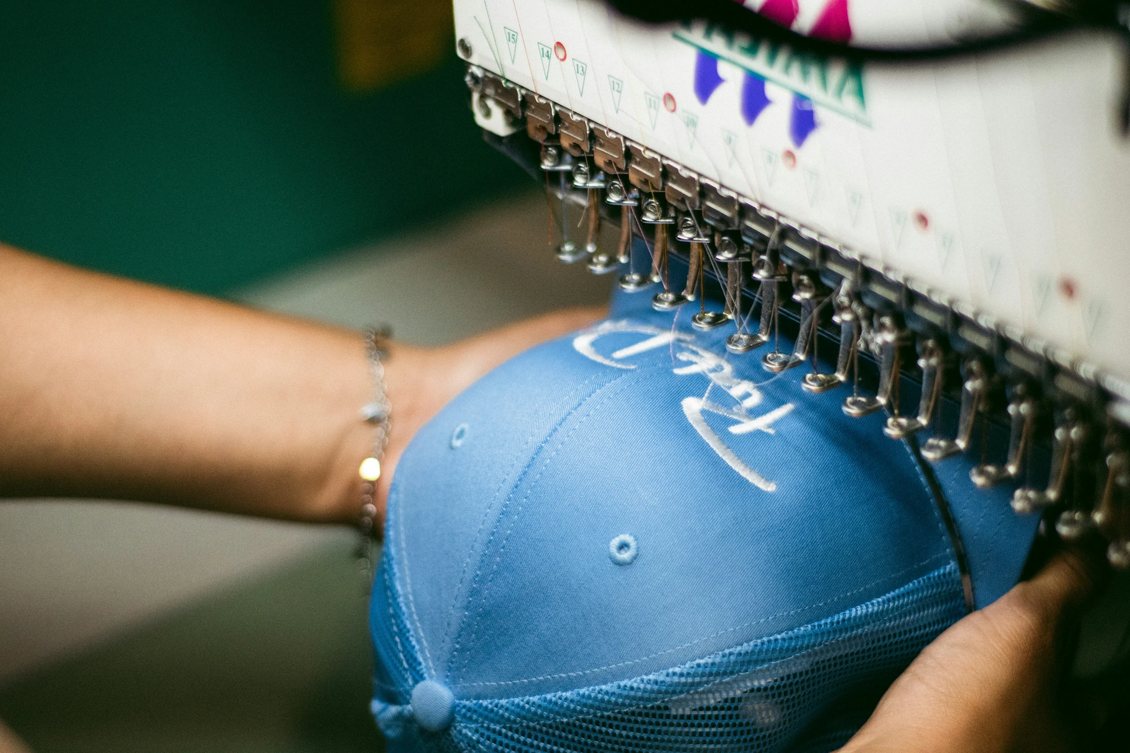 Una macchina da ricamo lavora un cappellino blu con scritta bianca; si vedono aghi, fili, tessuto e mani.