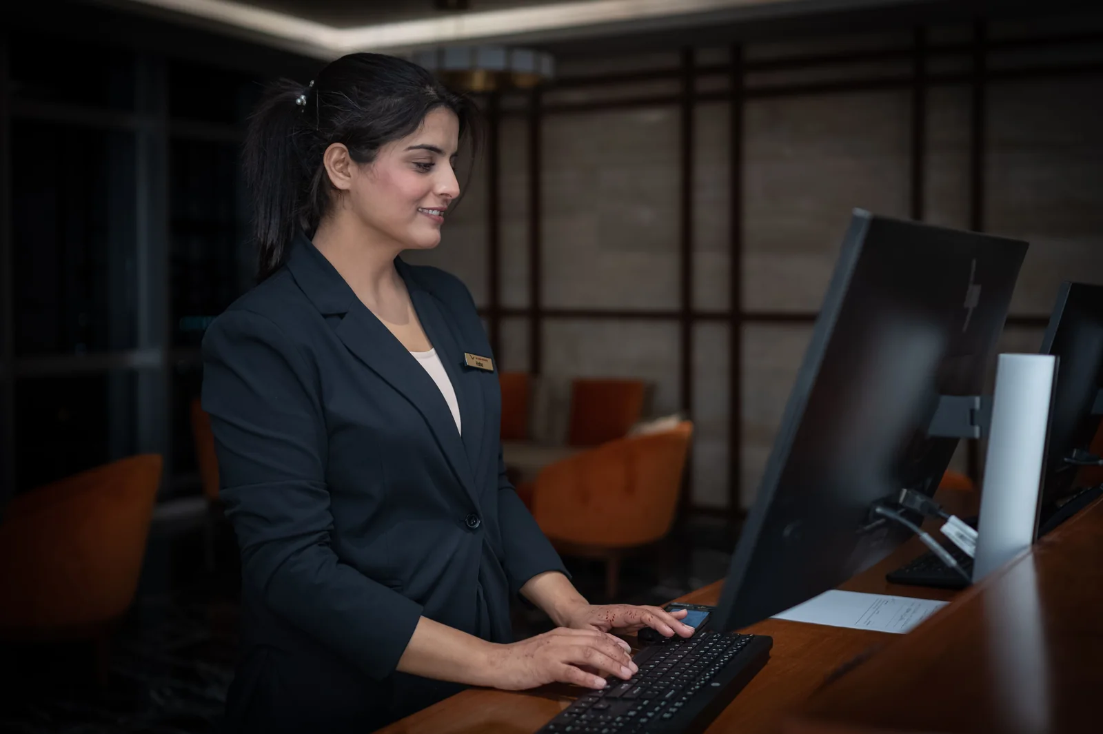 Una receptionist in giacca navy lavora al computer dietro il banco di un hotel.