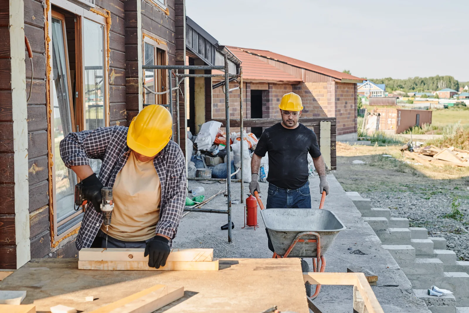 Due professionisti con casco giallo al lavoro in un cantiere, con trapano, legno e carriola.