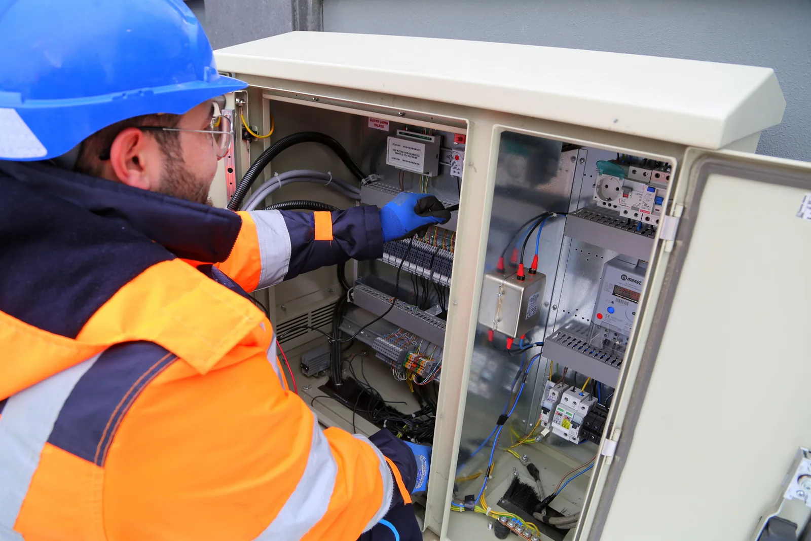 Tecnico in giacca arancione e navy mentre lavora su un quadro elettrico.