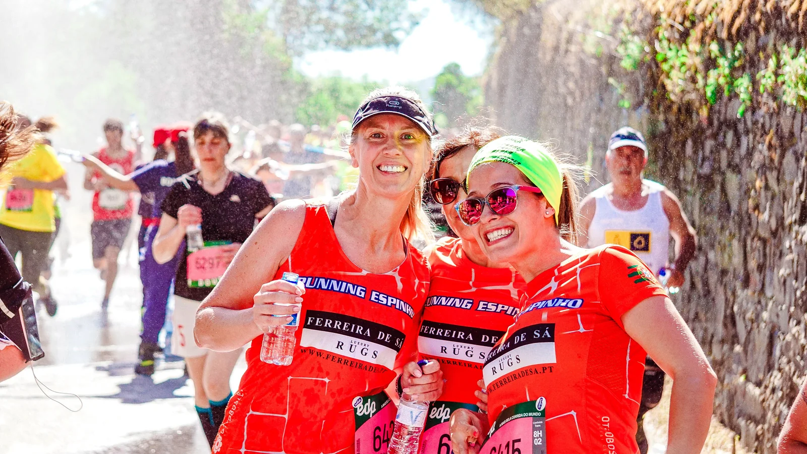 Tre partecipanti a una corsa sorridono insieme, con maglie arancioni e spruzzi d’acqua in controluce.